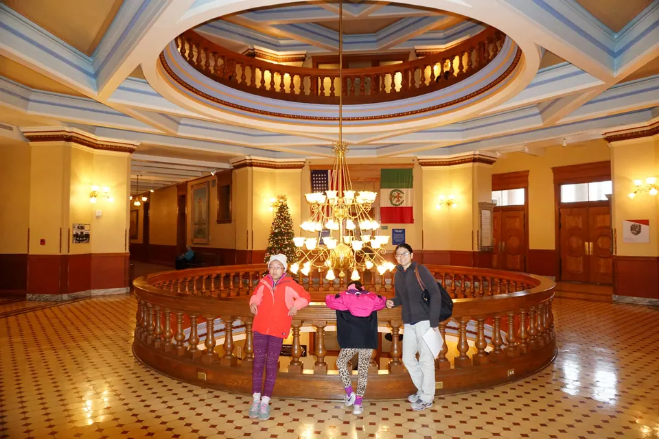 A woman and two girls lean on a round wooden railing under a chandelier, with a Christmas tree and flags on the far wall.