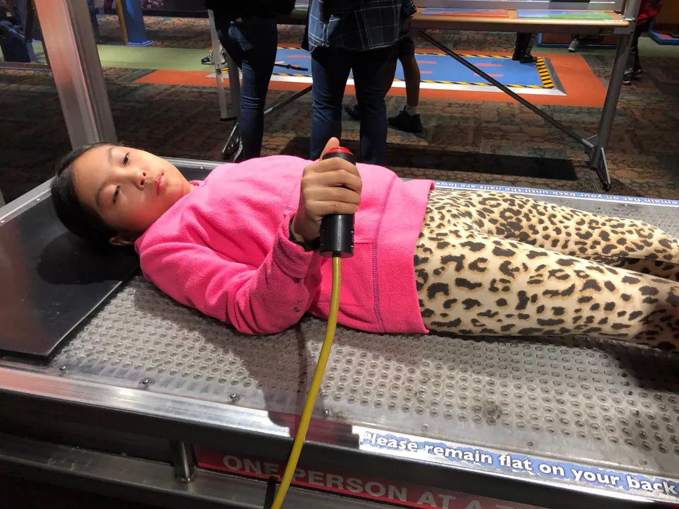A girl in a pink fleece and leopard-print leggings lies flat on a bed-of-nails exhibit, holding a control handle.
