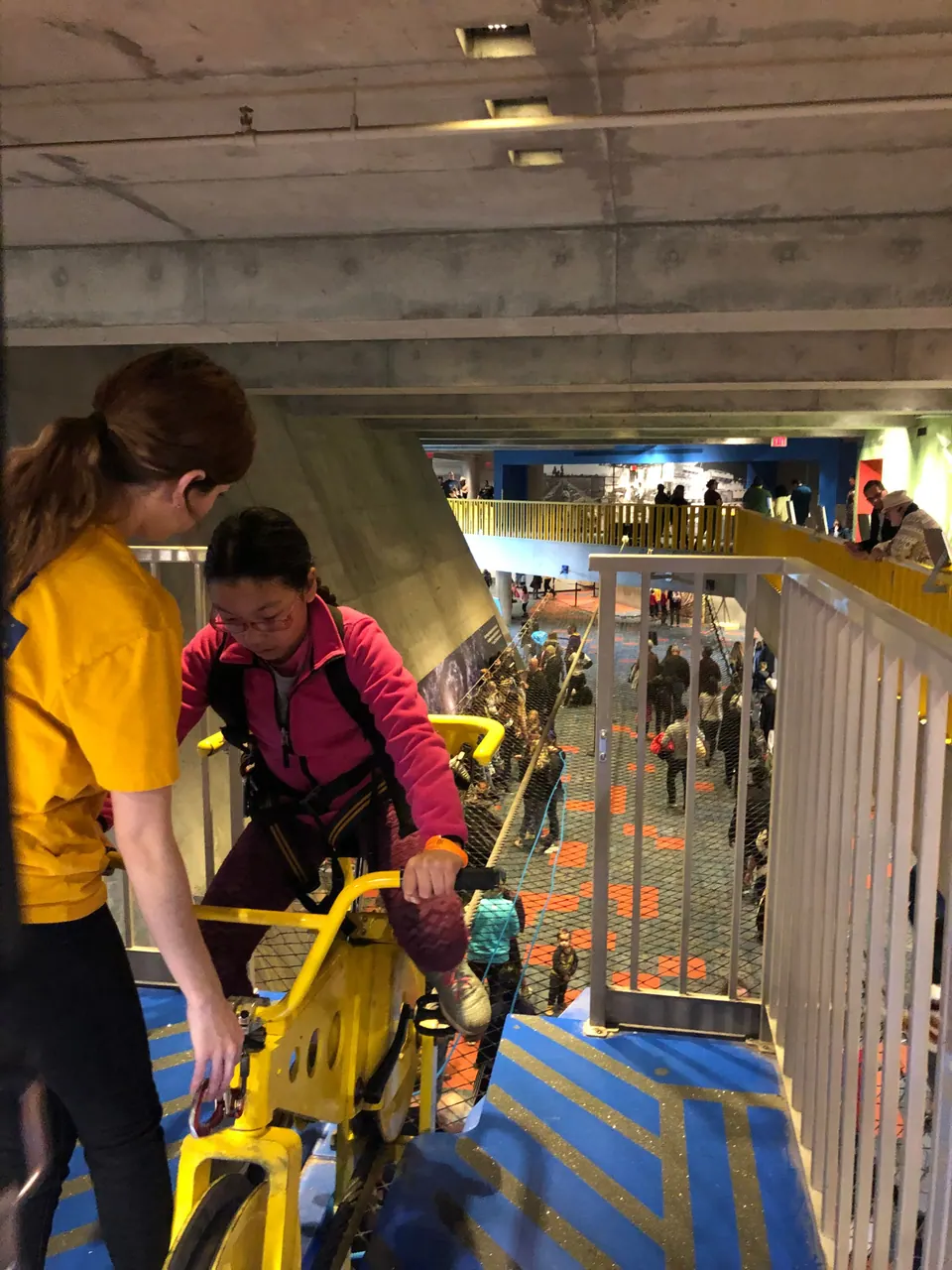 A girl in a harness sits on a yellow bicycle at the start of a high wire while a staff member in yellow checks her in; the busy floor is far below.