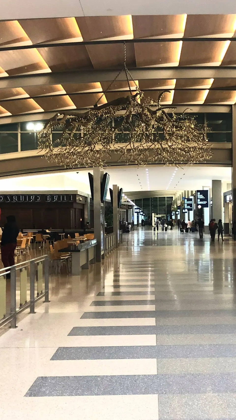A long, nearly empty airport concourse with a striped floor and a branching chandelier of small lights hanging from a wood-slatted ceiling.