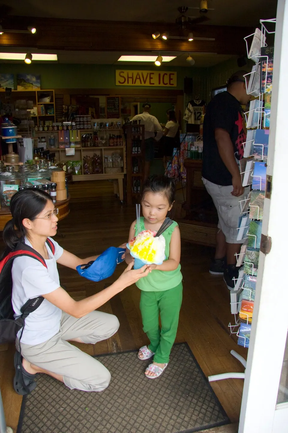 A woman with a backpack kneels in a shop doorway, handing a bag to a small girl in green who holds a shave ice; a Shave Ice sign hangs inside.