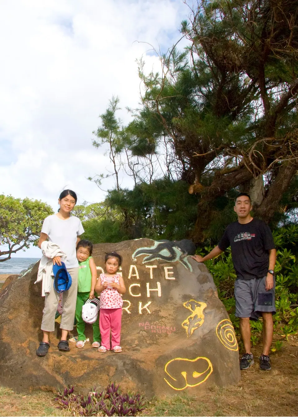 Two adults and two small girls stand around a large boulder painted with the words Lydgate Beach Park and yellow petroglyph shapes, under a windblown tree.