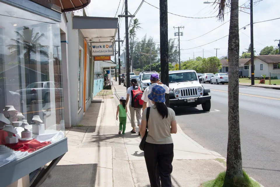 A woman with a backpack and a blue cap and a small girl in green walk along a shop-lined sidewalk with palm trees, a Jeep parked at the curb.