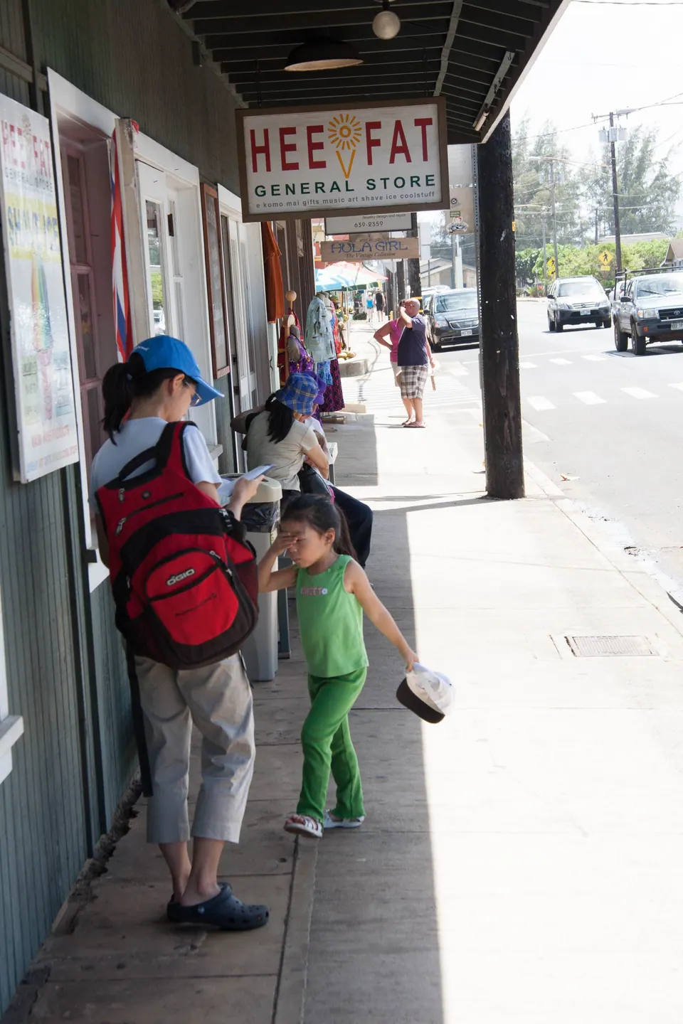 A woman with a red backpack and a small girl in green stand under a covered sidewalk beside a sign reading Hee Fat General Store.