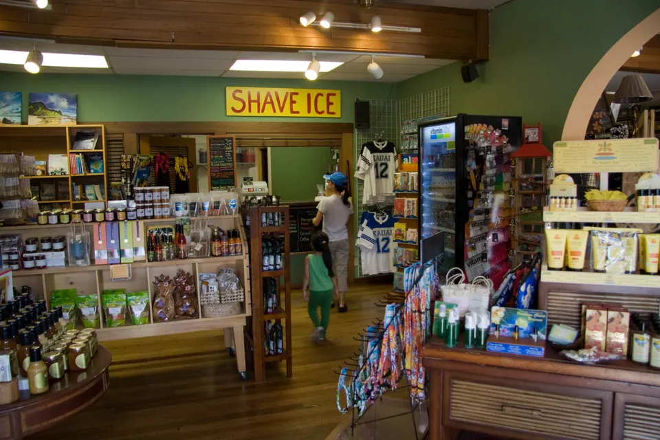 The shop interior: a yellow Shave Ice sign on a green wall, shelves of goods and postcards, and a small girl in green in the aisle.