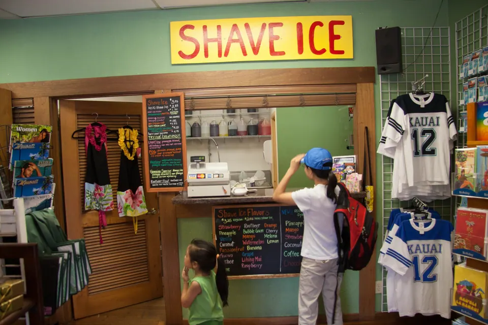 A woman with a red backpack and a blue cap orders at the counter under the Shave Ice sign while a small girl waits beside jerseys reading Kauai 12.