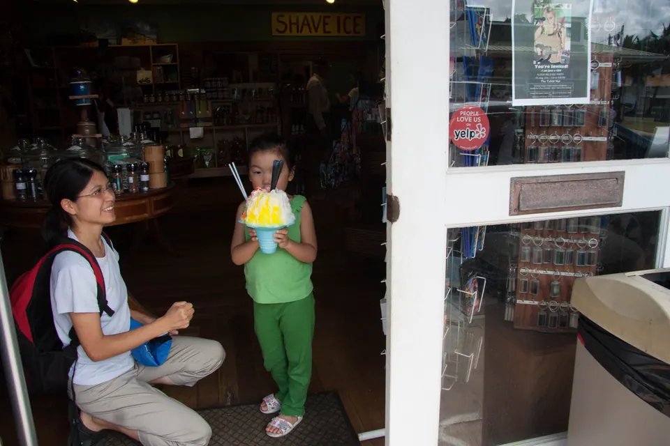 A woman kneels in the shop doorway with a red backpack while a small girl in green stands holding a big yellow-and-white shave ice.