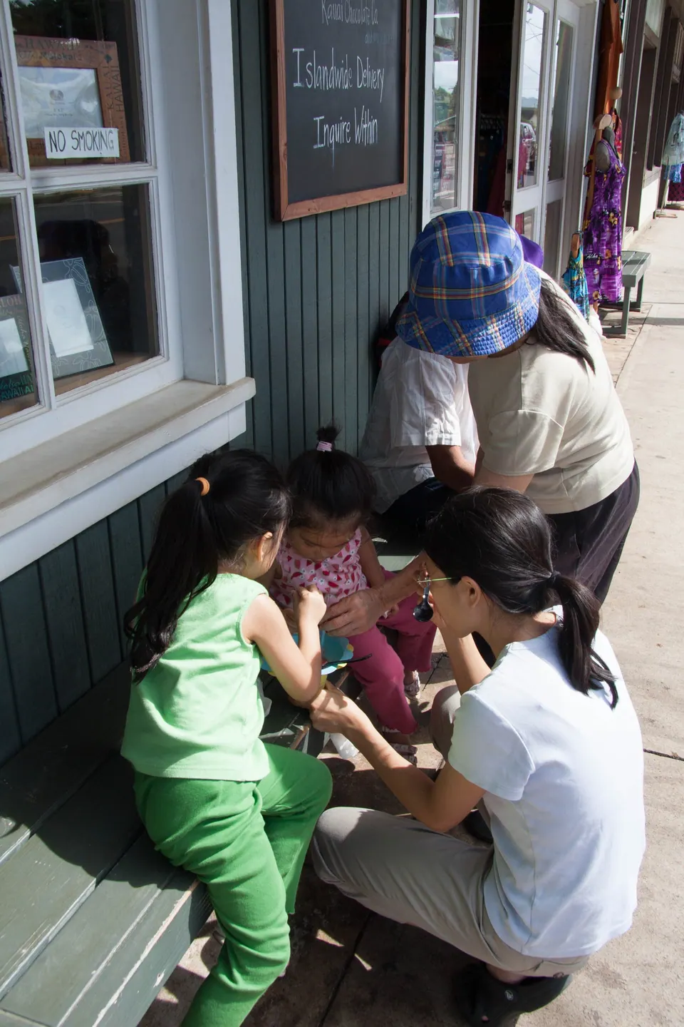 A small girl in green and a woman crouch together on the shop porch while another adult in a blue hat leans over them.