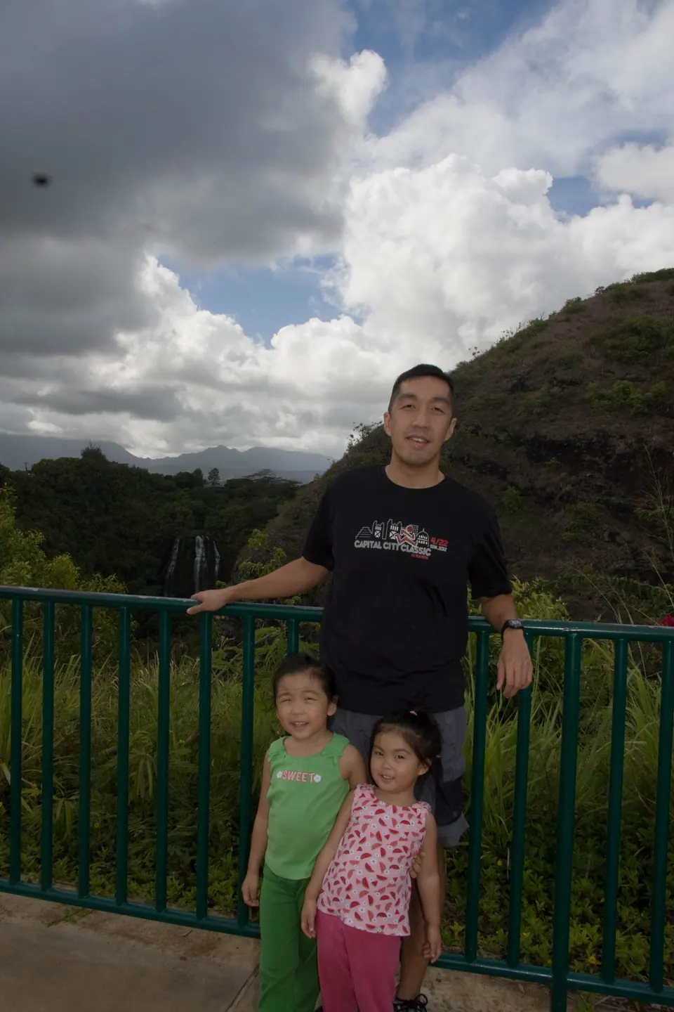 A man in a black tee leans on a green railing with two small girls in front of him, big clouds overhead and the falls just visible in the trees behind.