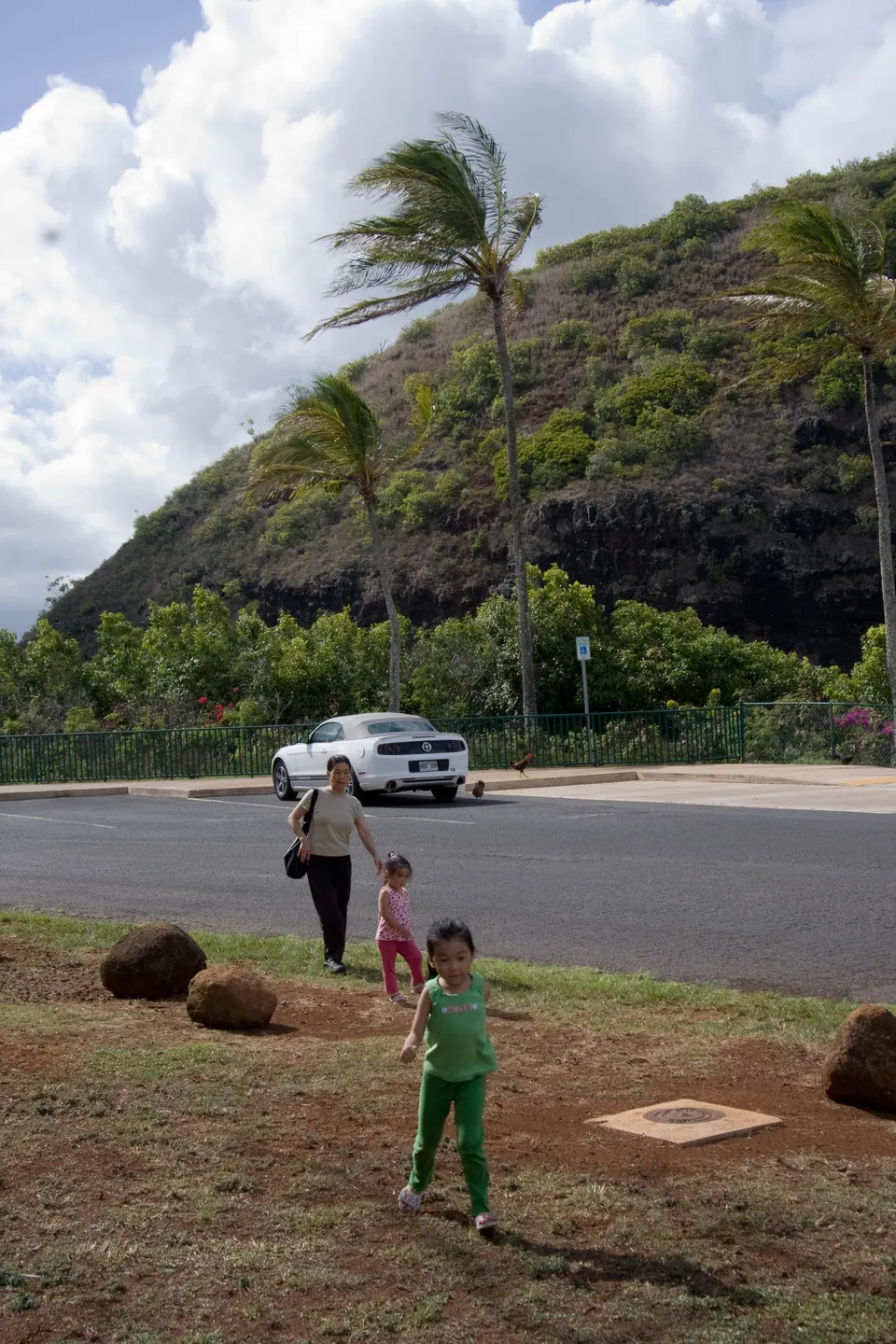 A green hill and palms above a parking area; a small girl in green walks across the lot toward a white vehicle.