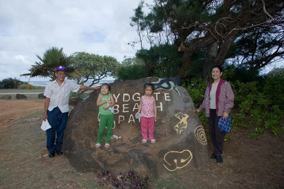 An older man and woman stand with two small girls, in green and pink, beside the big boulder painted Lydgate Beach Park under a windblown tree.