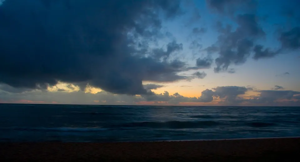 Dark storm clouds over the sea with a thin band of gold sunrise light on the horizon and dim sand in the foreground.