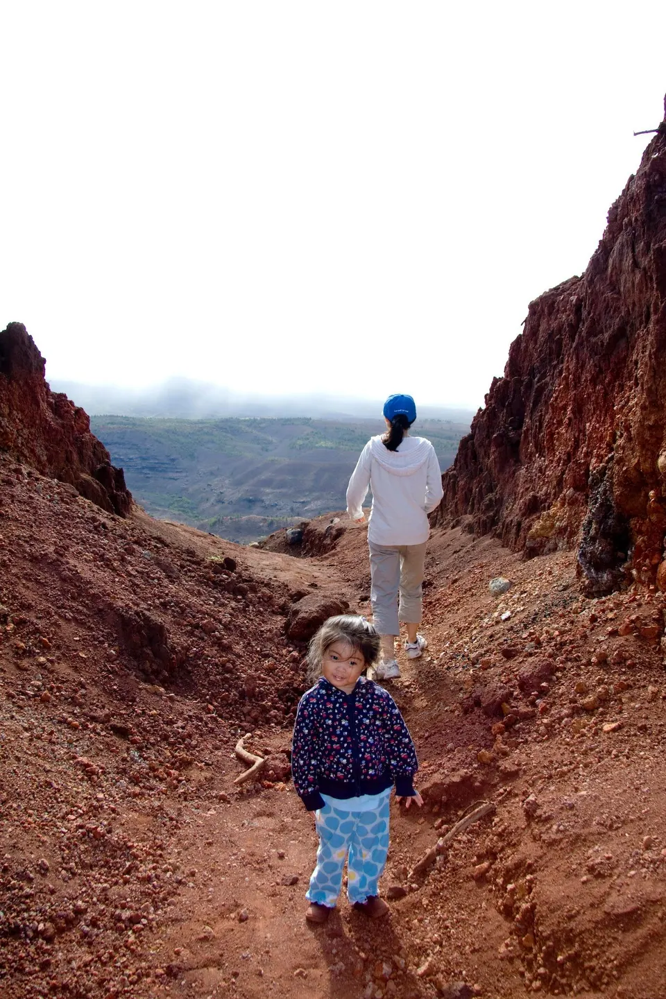 A small girl in a purple jacket stands on a narrow path between eroded red banks; an adult in a white jacket and blue cap walks ahead toward a hazy valley.