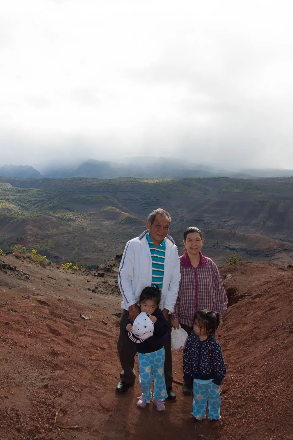 An older man and woman stand with two small girls in matching blue patterned pants on a red-dirt ridge, with green hills and low cloud behind.