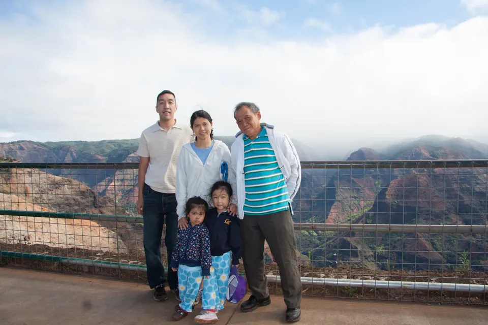 Three adults and two small girls stand at a metal railing; behind them, layered red canyon walls disappear into cloud.