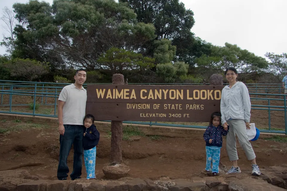 Two adults and two small girls stand on either side of a brown wooden sign reading Waimea Canyon Lookout, Division of State Parks, Elevation 3400 ft.