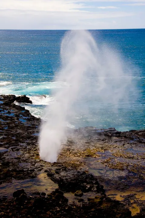 A tall plume of white spray bursts from a hole in black lava rock, with deep blue ocean behind.
