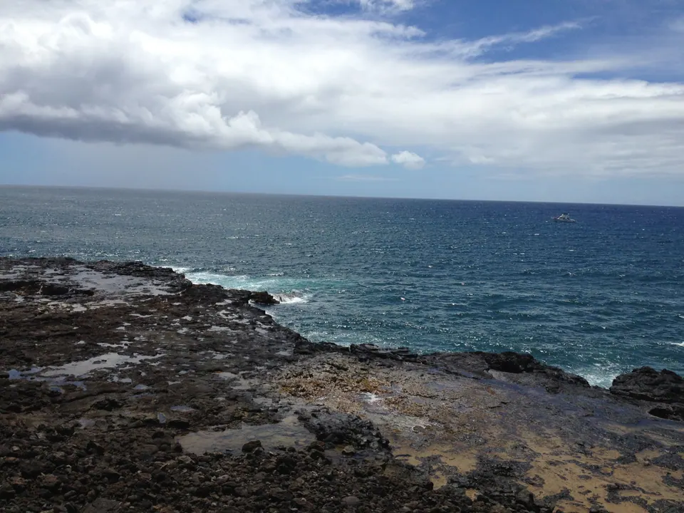 A black lava shelf with tide pools meeting the deep blue sea under big clouds, a boat far out on the water.