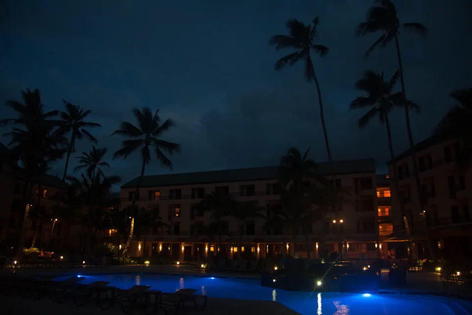 A multi-storey resort building with lit windows behind a dark garden, tall palms against a dark sky, and the pool glowing blue at the edge of the frame.