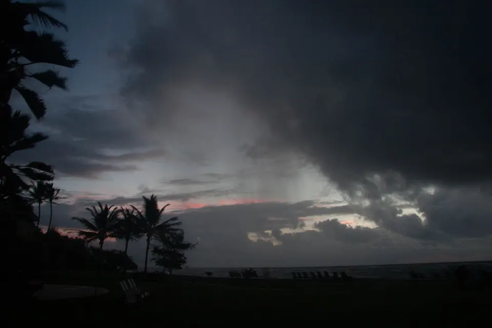 A stormy dawn sky with a dark cloud mass and a thin red glow low on the horizon behind palm silhouettes.