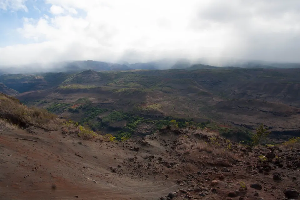 A broad view from red dirt across dry brown-green canyon country, with cloud lying along the far rim.
