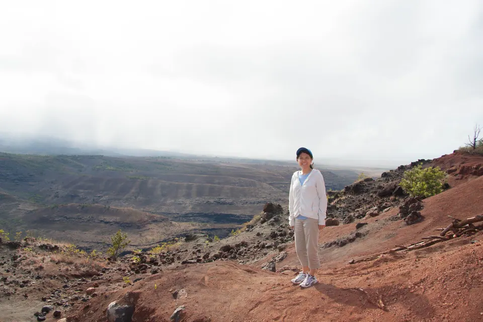 An adult in a white jacket stands alone on the red-dirt rim, the wide canyon and low cloud behind.