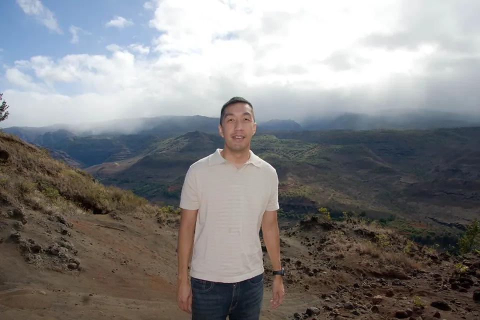 A man in a cream shirt stands on the red-dirt rim with the misty canyon behind him.