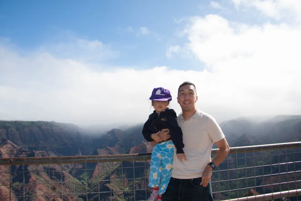 A man in a cream shirt holds a small girl in blue patterned pants at a railing, canyon and cloud behind.
