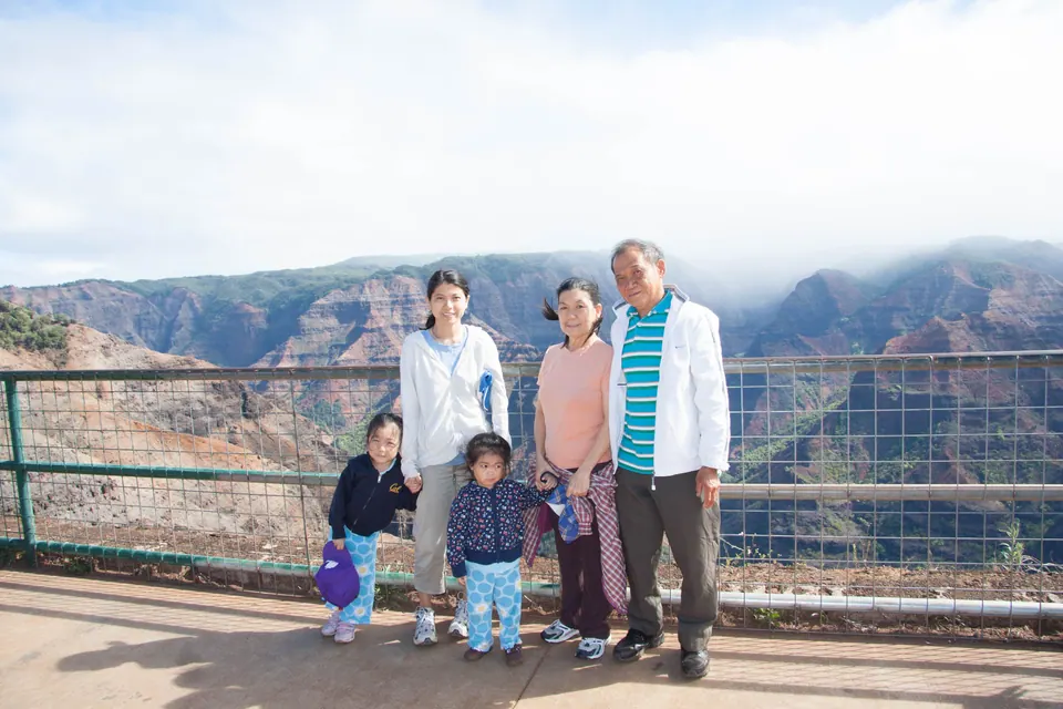 Adults and two small girls line up at a wire-mesh railing with layered red canyon walls and low cloud behind them.