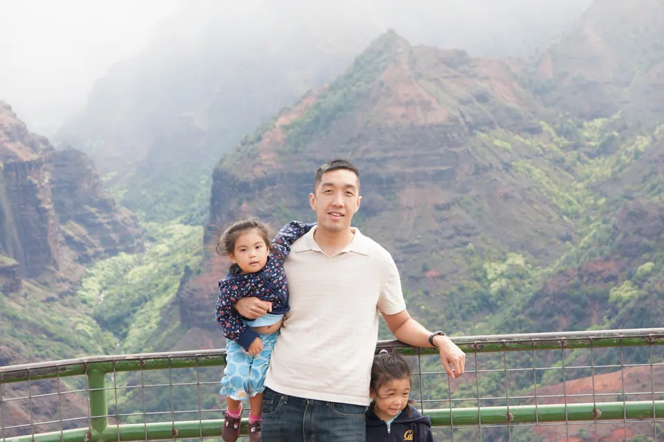 A man in a cream shirt with a small girl in blue on his arm leans on a green railing; steep mist-covered canyon walls behind.