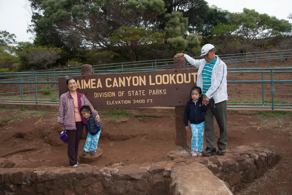 An older man and woman with two small girls stand at the brown Waimea Canyon Lookout sign, red dirt underfoot and pines behind.