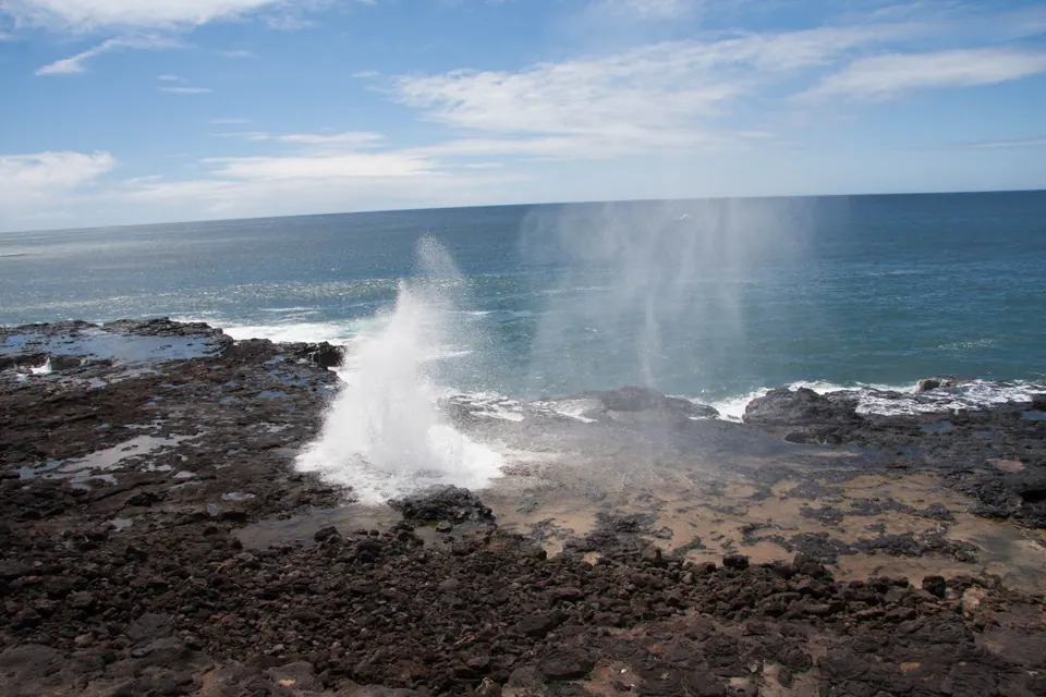 Spray bursts up from a black lava shelf in a broad view of ocean and blue sky.