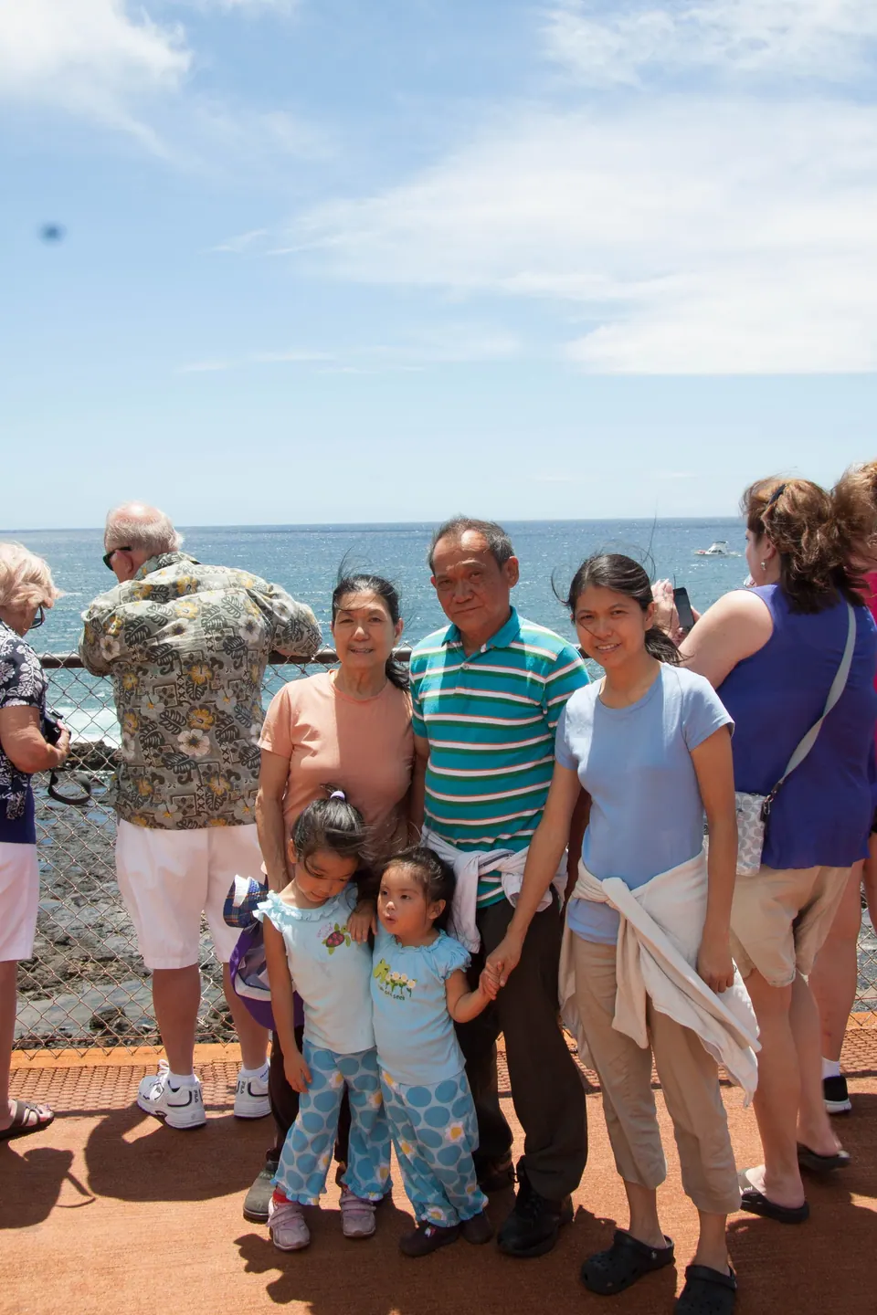 An older man and woman, a younger woman and two small girls pose on an orange-paved viewing platform at a sea railing, other visitors behind.
