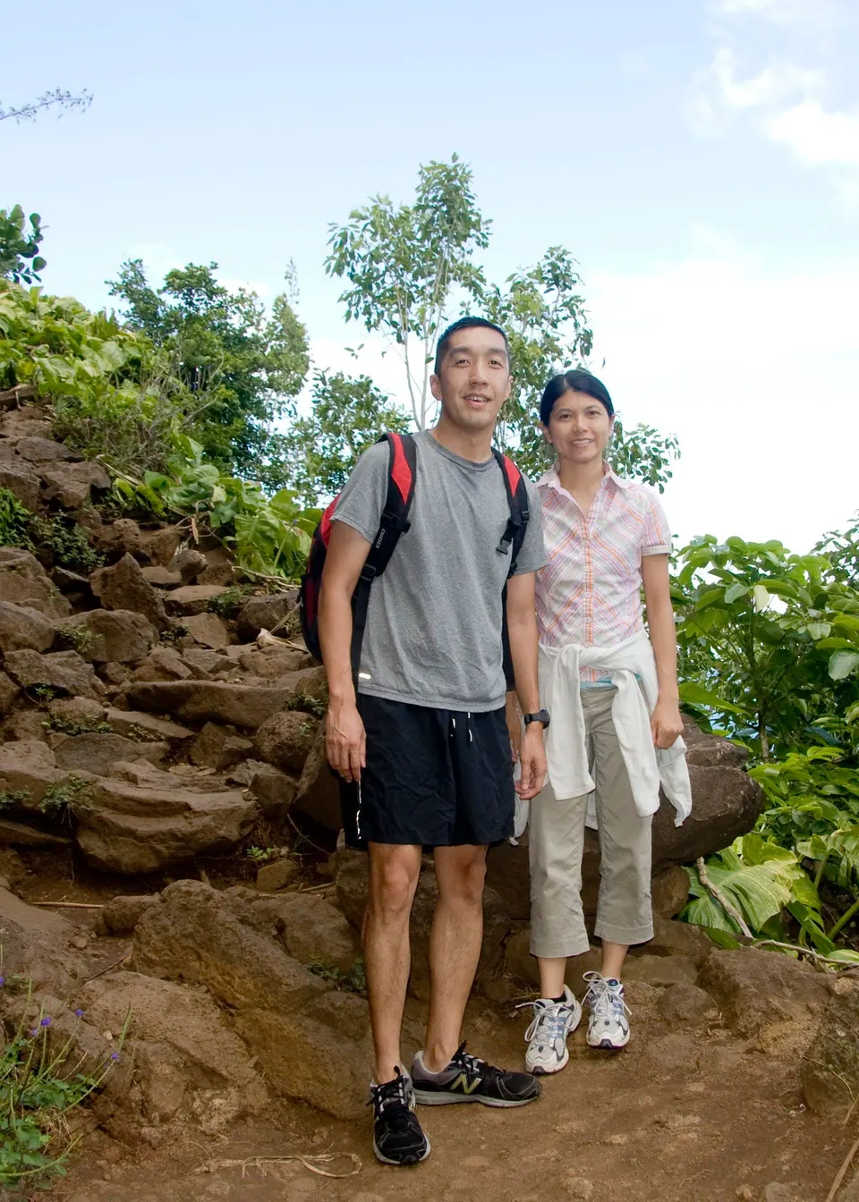 A man with a backpack and a woman in a pink top stand on a rocky, root-covered path through jungle.