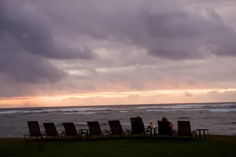 Purple clouds and a pink glow over the sea, a row of empty lounge chairs on the dark lawn.