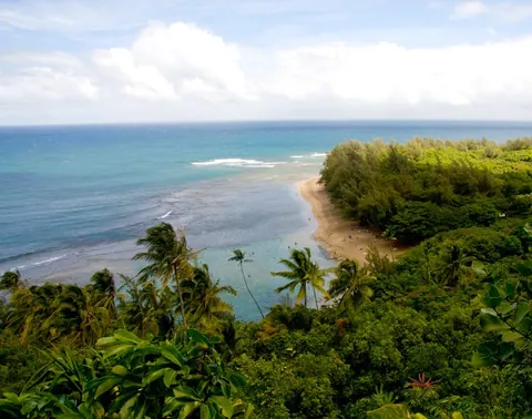 A wide view over jungle and palms to a curving strip of white sand and turquoise reef water under big clouds.