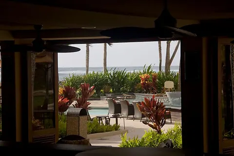 An open-air lobby with ceiling fans looking out to a patio with red tropical plants, chairs and the ocean beyond.