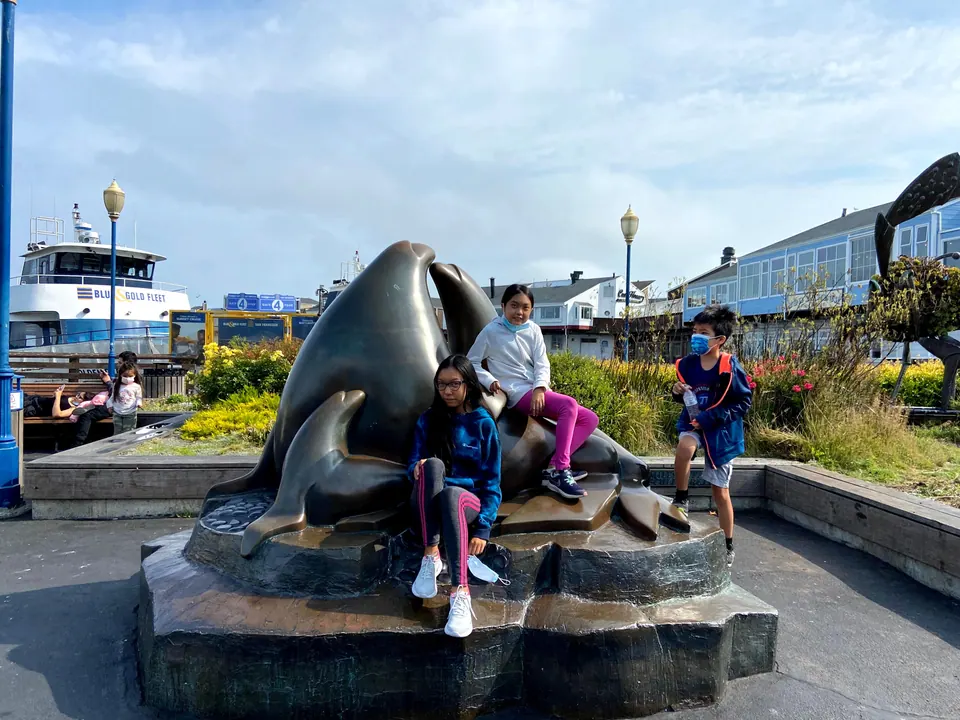 Two girls and a masked boy in a blue jacket sit and stand on a bronze sculpture of sea lions, with a Blue and Gold Fleet boat and blue shopfronts behind.