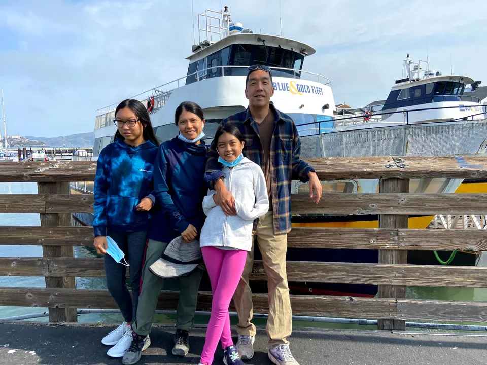 Four people pose at a wooden railing in front of a white ferry, one girl holding her mask, with the bay and hills behind.