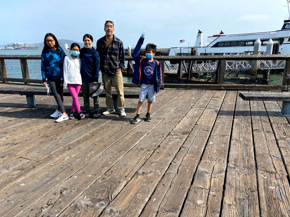 Five people stand in a row on weathered pier boards, a boy in a blue jacket raising a water bottle, with Alcatraz Island and the hills across the bay behind.