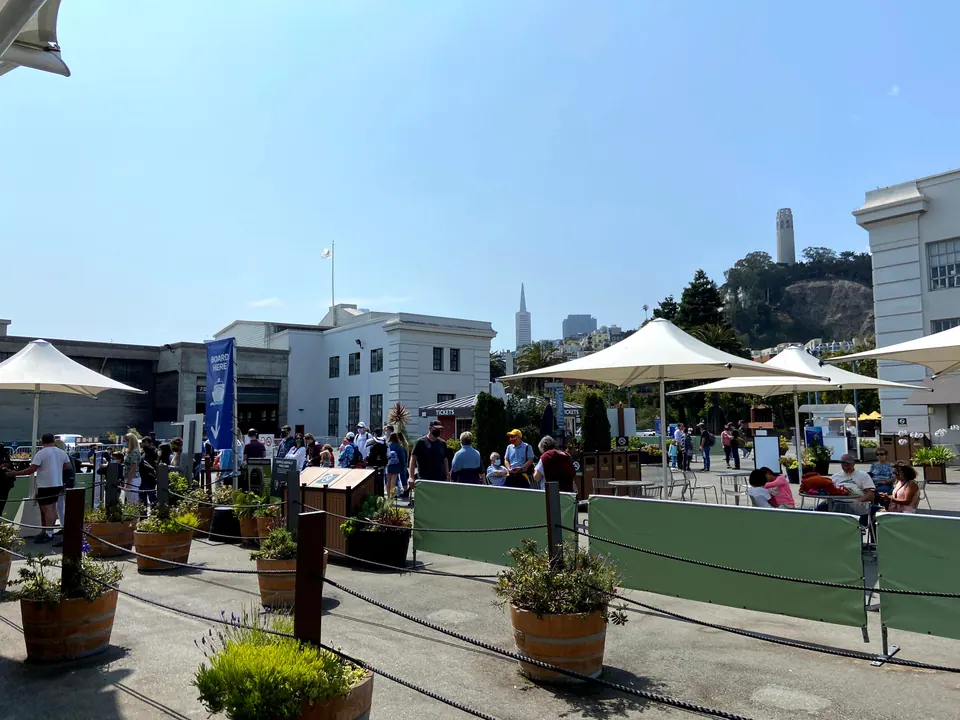 A courtyard with a blue Board Here banner, roped queue lines and white umbrellas, and Coit Tower and a pointed skyscraper above the trees.