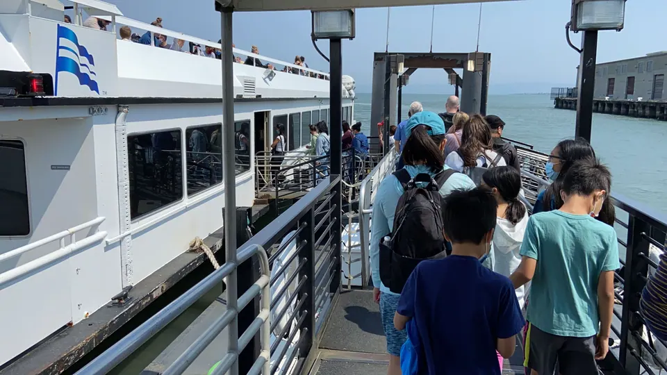 Passengers in masks file down a gangway toward a white ferry with a blue wave logo, with the bay beyond.
