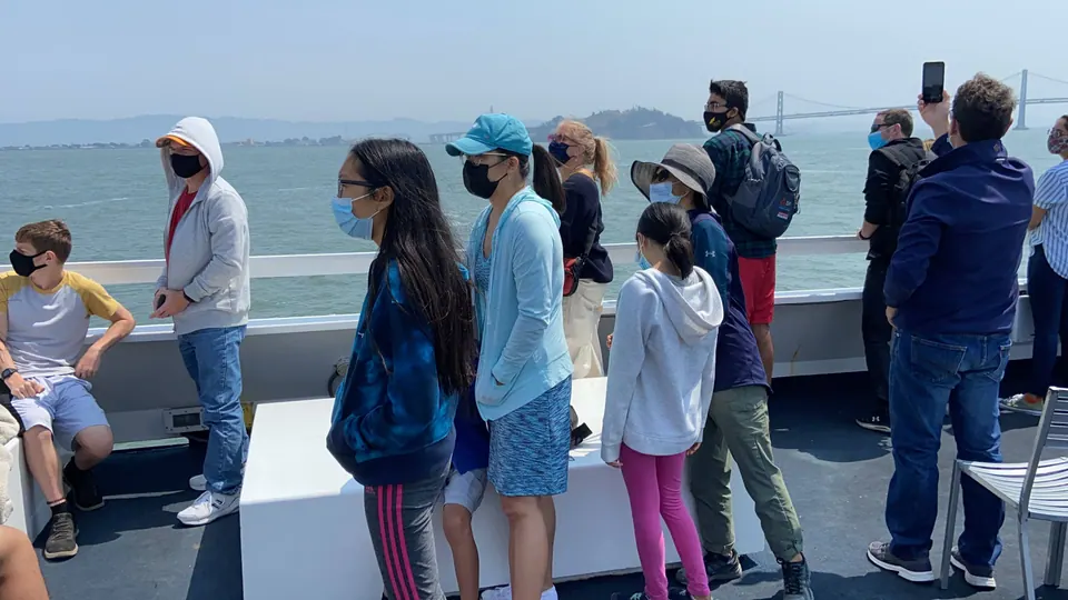 Passengers at a ferry rail look toward a long bridge and a green island across hazy water, two girls among them in masks and, between them, a woman in a teal cap and a light blue jacket.