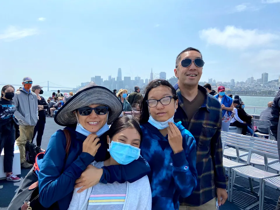 Four people crowd together on an open ferry deck, one hugging another, with a hazy city skyline and bridge behind them.