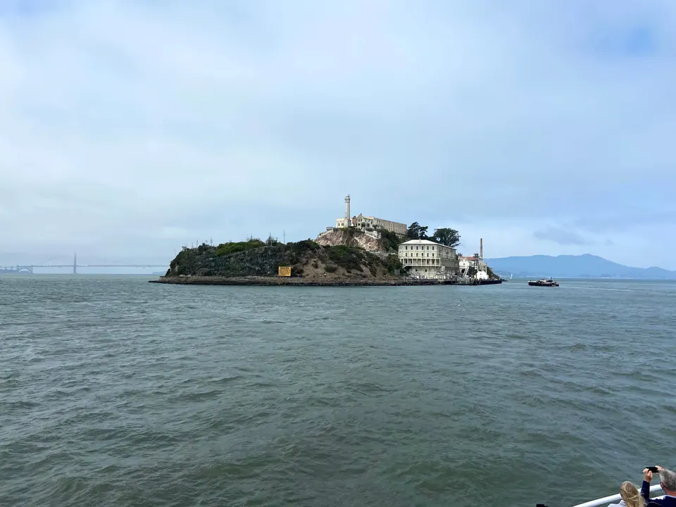 Alcatraz Island across grey water from the approaching ferry, with the lighthouse and cellhouse on top, the Golden Gate Bridge faint in the haze at left and a small boat at right.