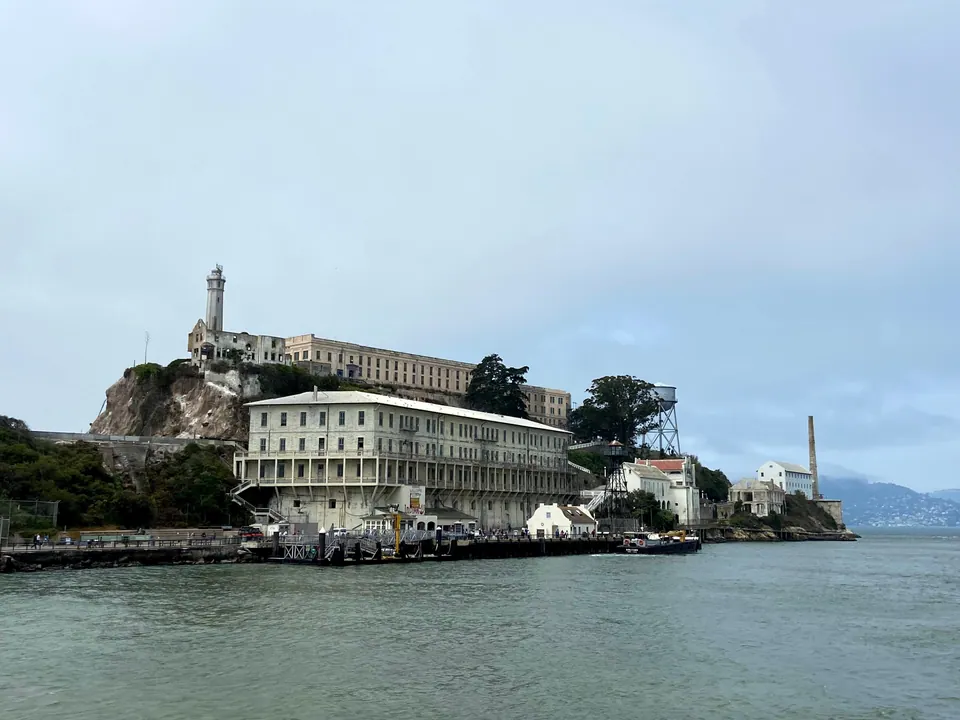 A pale cellhouse and a dock below a lighthouse and water tower on an island, under a low grey sky.