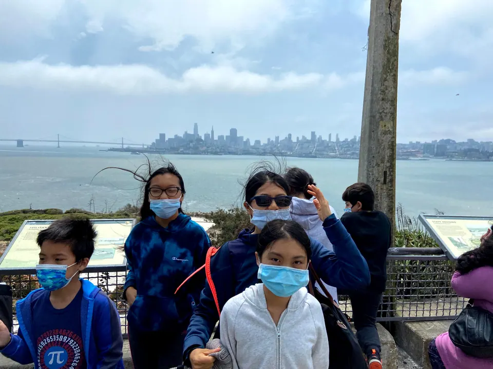 Wind-blown masked faces, including a boy in a blue jacket at left, crowd the foreground at a viewing rail beside two information signs, with a city skyline and a bridge hazy across the water.