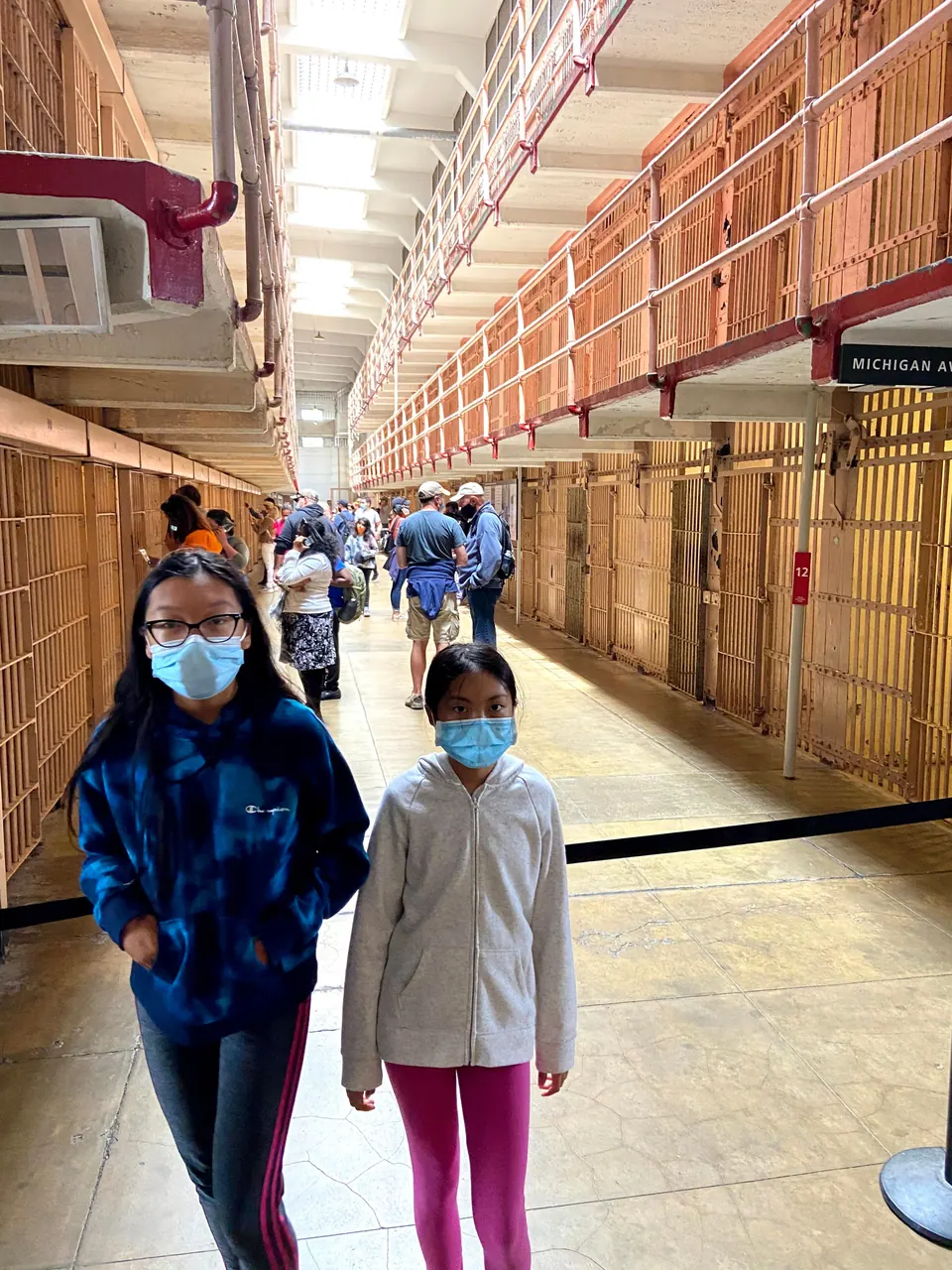 Two masked girls stand between tall tiers of cells, with other visitors further down the corridor and a stanchion rope at the right.