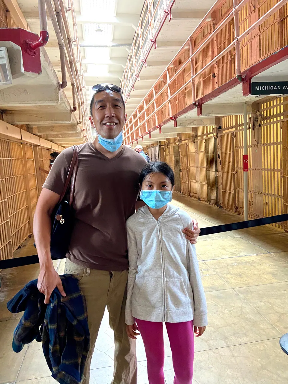 A smiling man with sunglasses on his head and his mask pulled down puts an arm around a masked girl in front of tiers of cells.