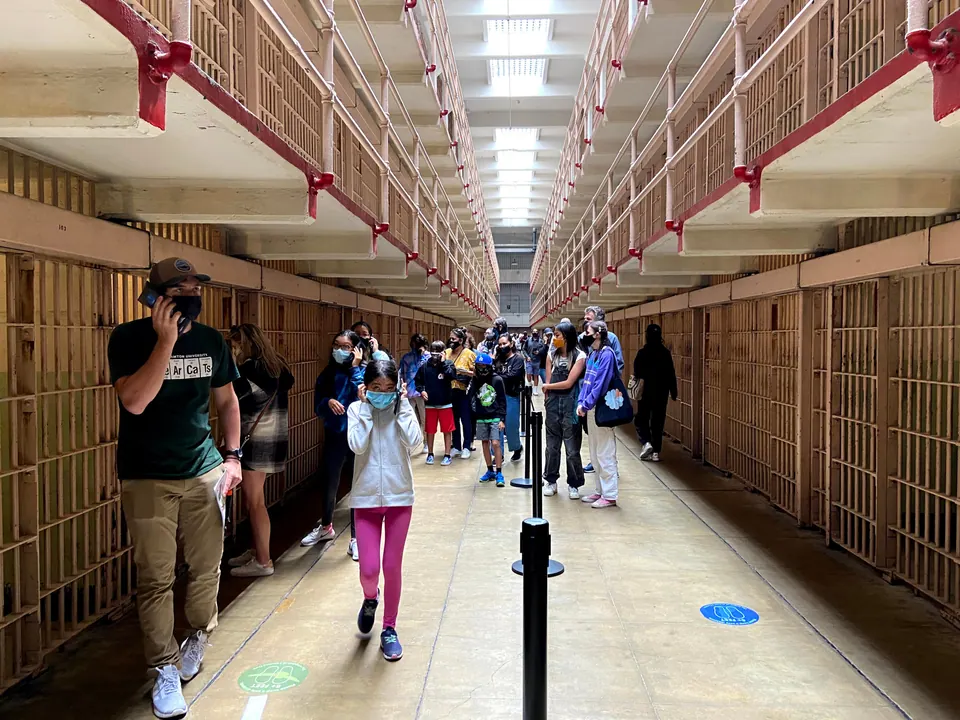 Visitors in masks line up with audio-tour phones in a long corridor between two tiers of cells, with distancing markers on the floor.